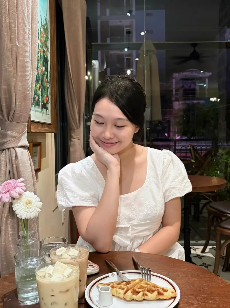 Eyes closed, chin on her hand, in a white eyelet dress beside iced lattes and gerberas.