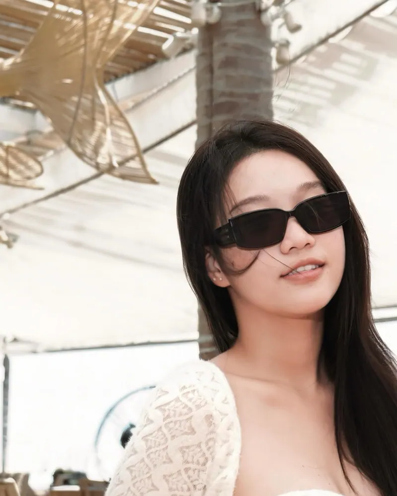 Bảo Ngọc by day: ivory lace, long dark hair and black rectangle sunglasses under a woven café canopy.