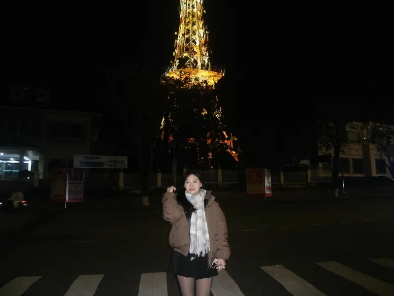 Standing on a crosswalk at night in a brown puffer, the tower lit above her.
