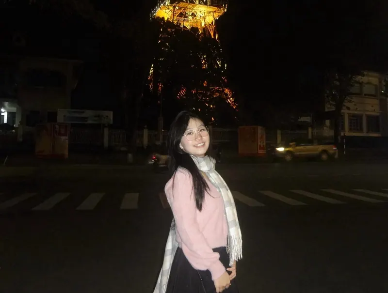 Laughing in a plaid scarf at night, a lit lattice tower behind her.