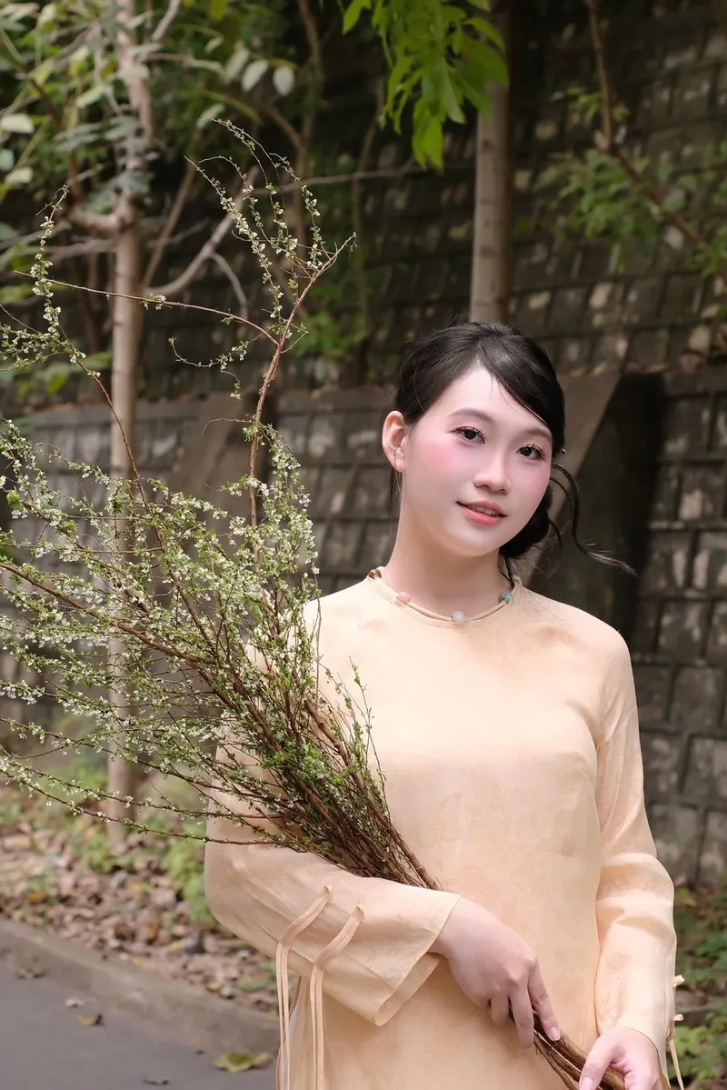 Look 01: an apricot silk áo dài with tie sleeves, hair up, a branch of white blossom in her arms.