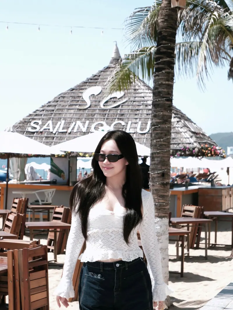 Look 04: an ivory lace top with dark denim and sunglasses, walking past a thatched beach bar.