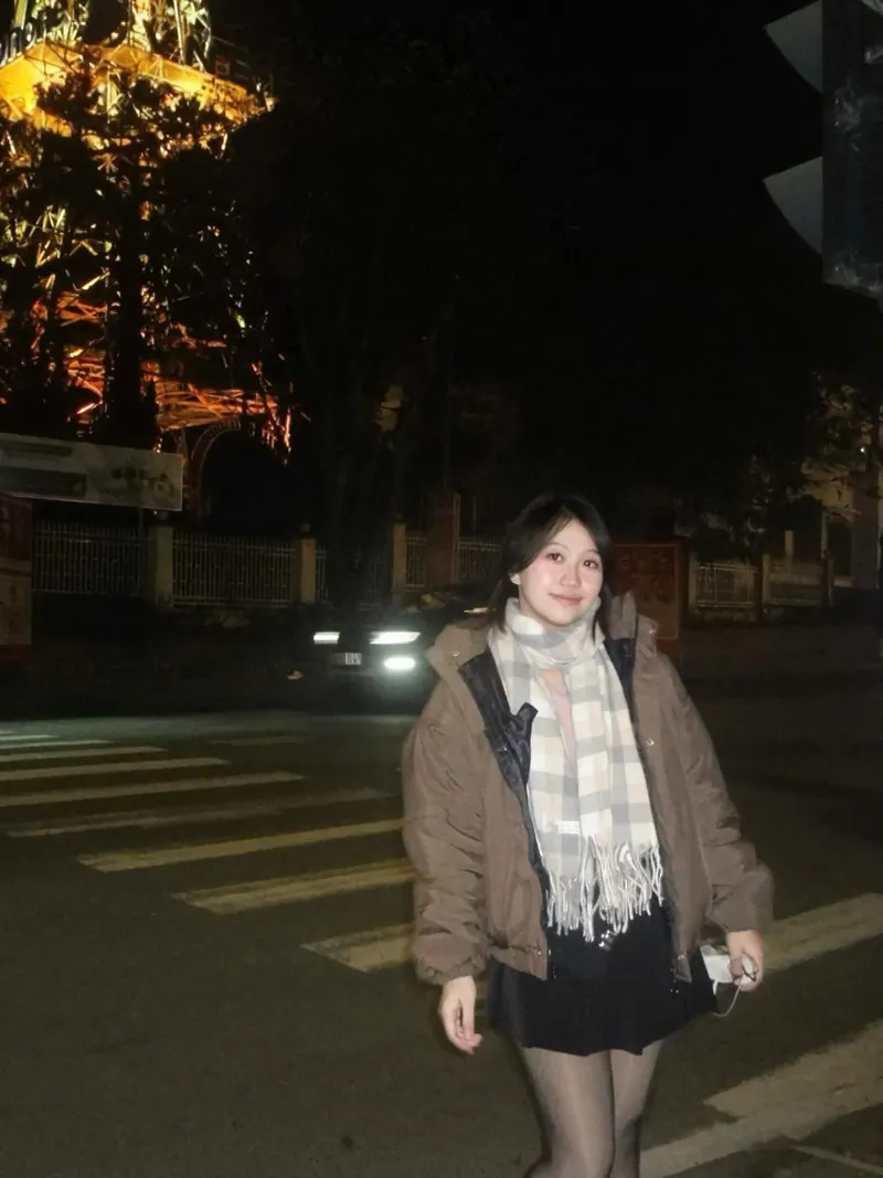 Look 06: a brown puffer, a plaid scarf, a black mini and sheer tights on a crosswalk at night, a lit tower behind her.