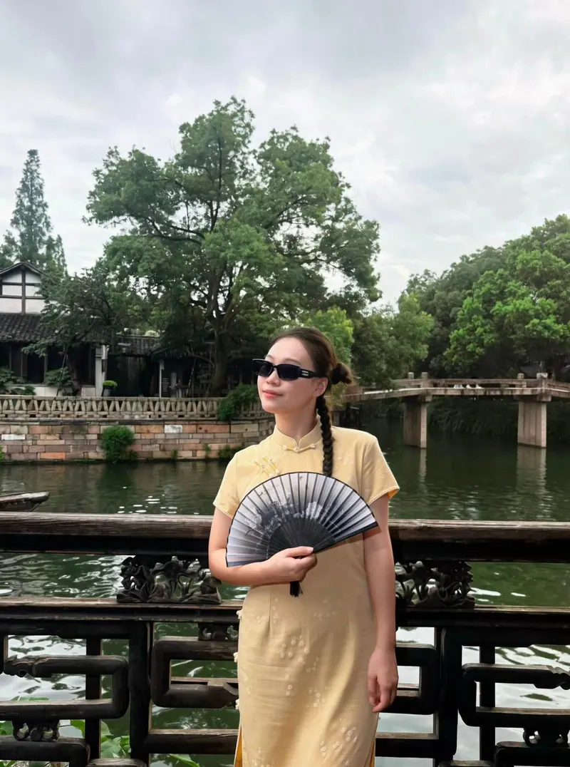 Look 05: a butter-yellow qipao, a single braid and an ink-painted fan beside a carved wooden railing by the water.
