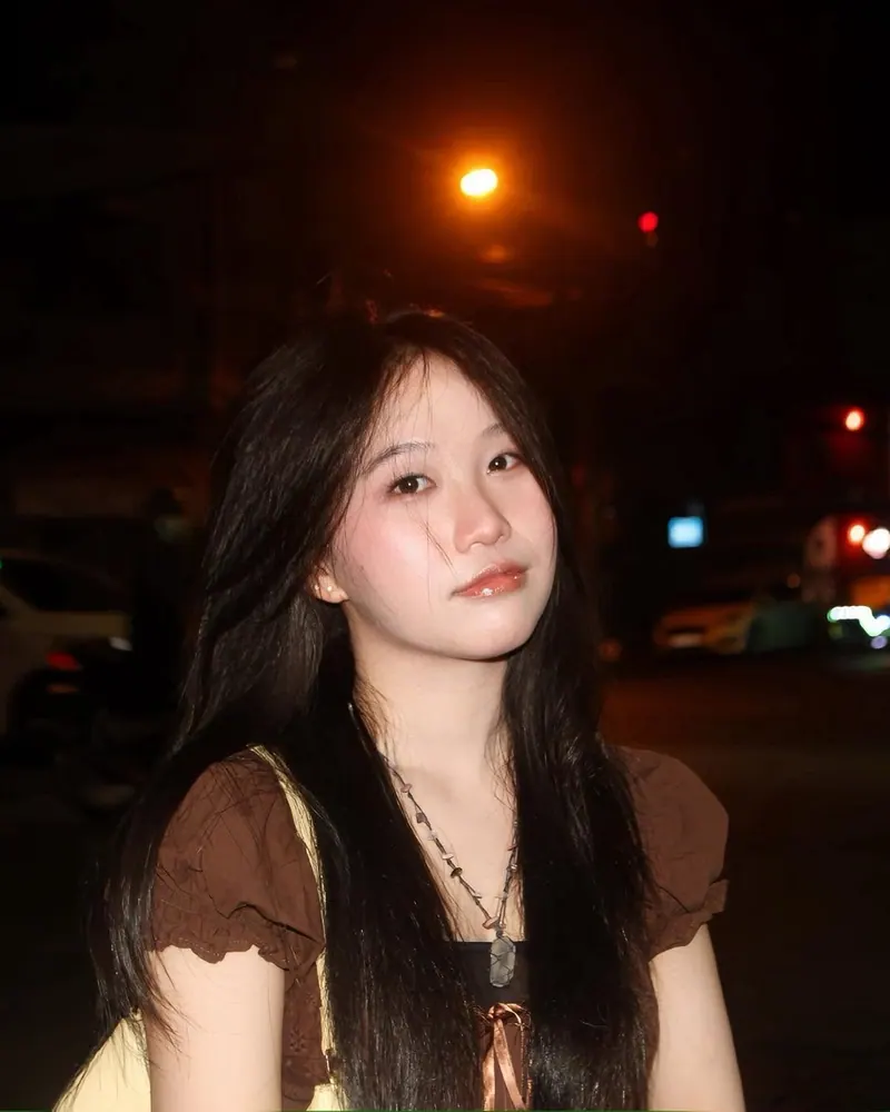 Bảo Ngọc after dark: a brown ruffled top and a stone pendant, lit by camera flash on a night street.