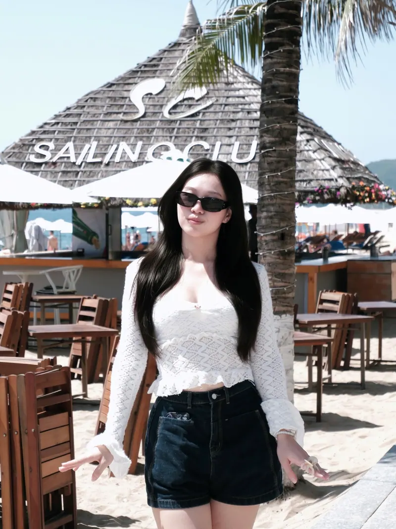 In ivory lace and denim beside a thatched beach bar, sunglasses on, a hand at her hair.