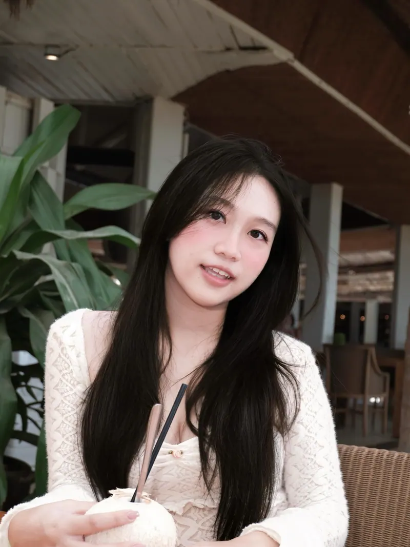 Bảo Ngọc in ivory lace on a shaded terrace, smiling softly beside a tropical plant.