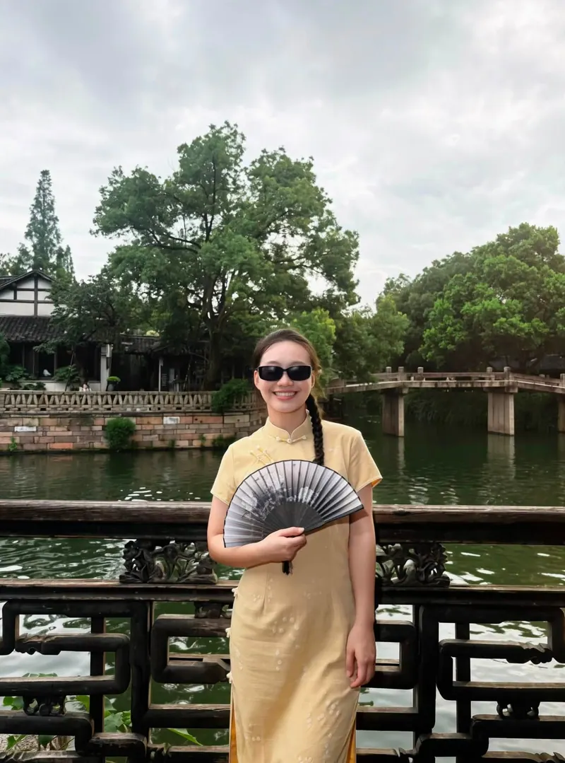 Smiling in a yellow qipao on a stone bridge, white-walled houses along the water.